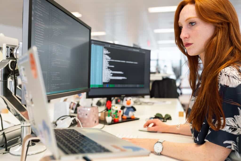 Woman sitting writing code on three computer screens Are Coding Bootcamps Worth It