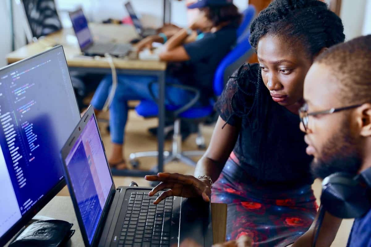 A picture of two people looking at a laptop placed next to a desktop, both displaying code. Coding Bootcamp vs College