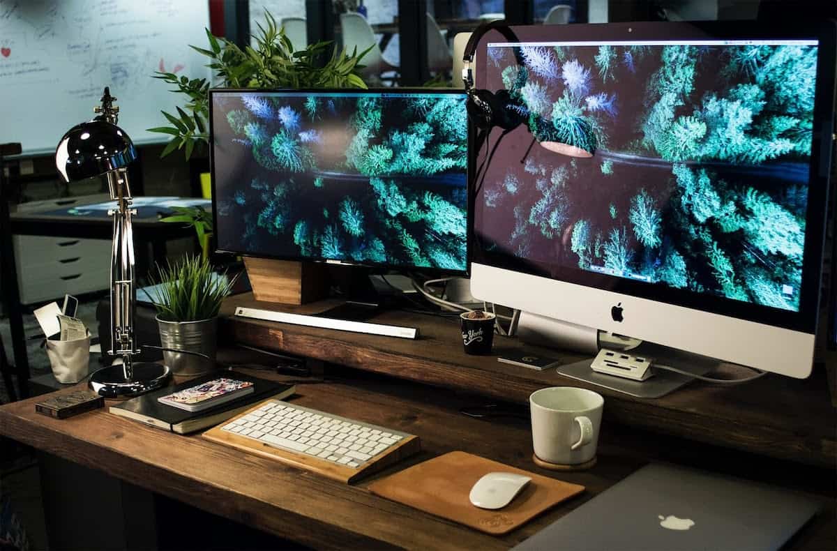 Monitors sitting on a desk with a cup, keyboard, and other things. Front End vs Backend Development