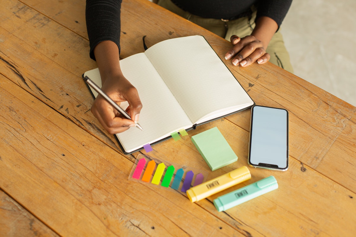A woman writing in a notebook, with a smartphone, highlighters, and sticky notes beside her on a wooden table