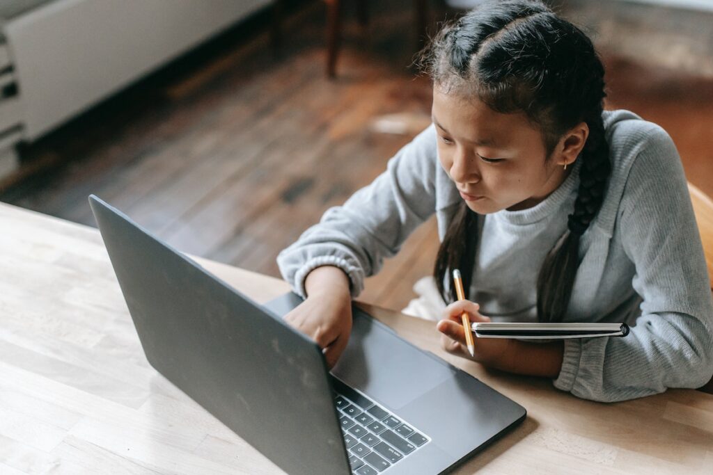 A girl typing on laptop and holding a pencil and notebook in the other hand