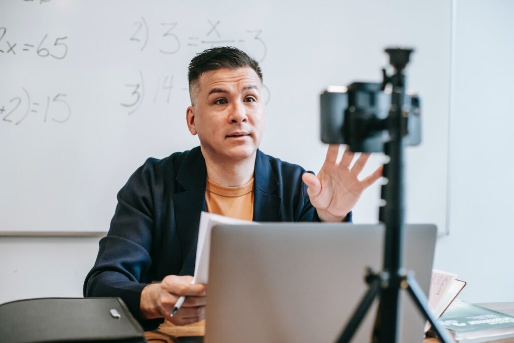 A teacher conducting an online class with a video camera and a laptop in a classroom environment.