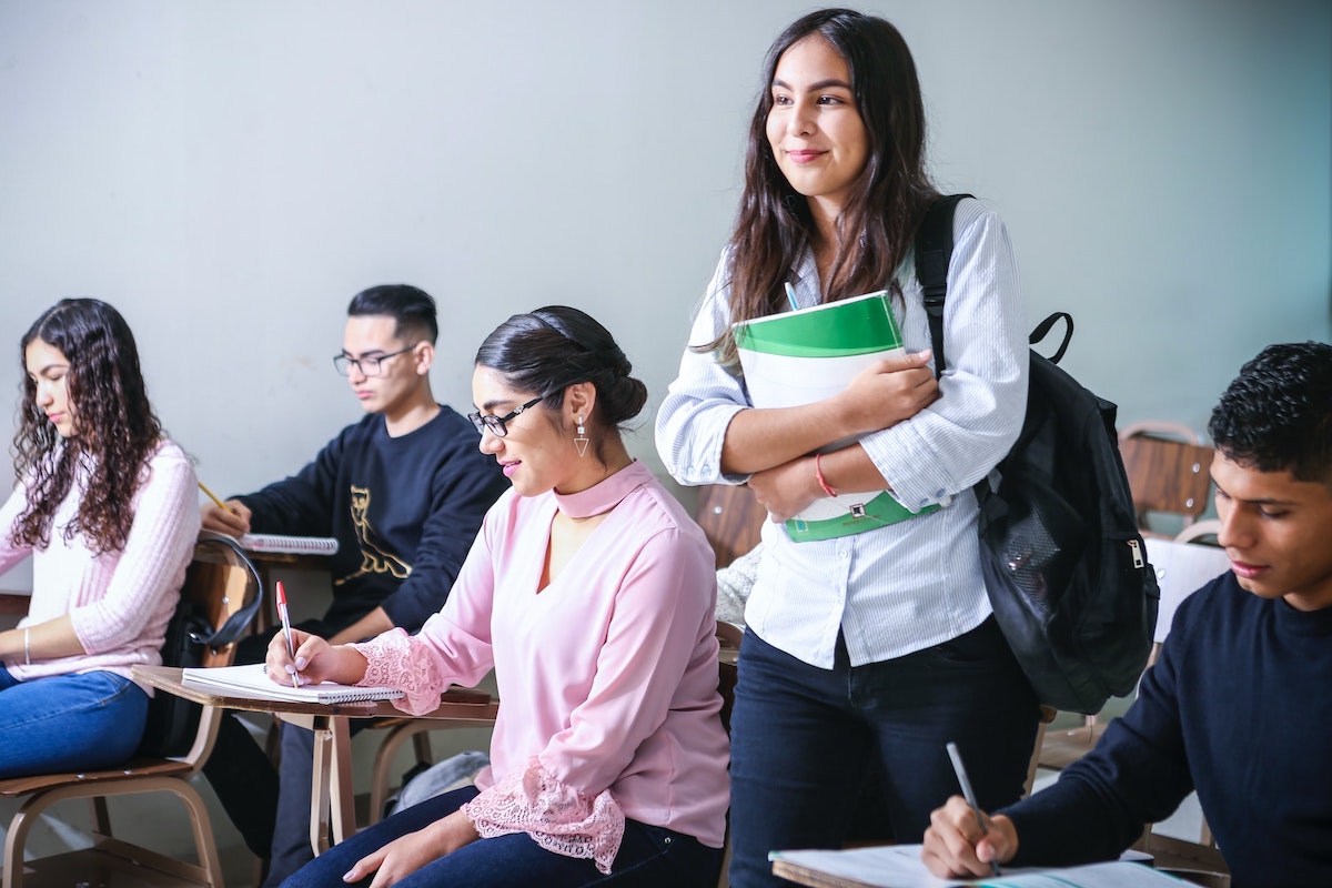 A group of students sitting at desks in a classroom and one student standing up holding a notebook