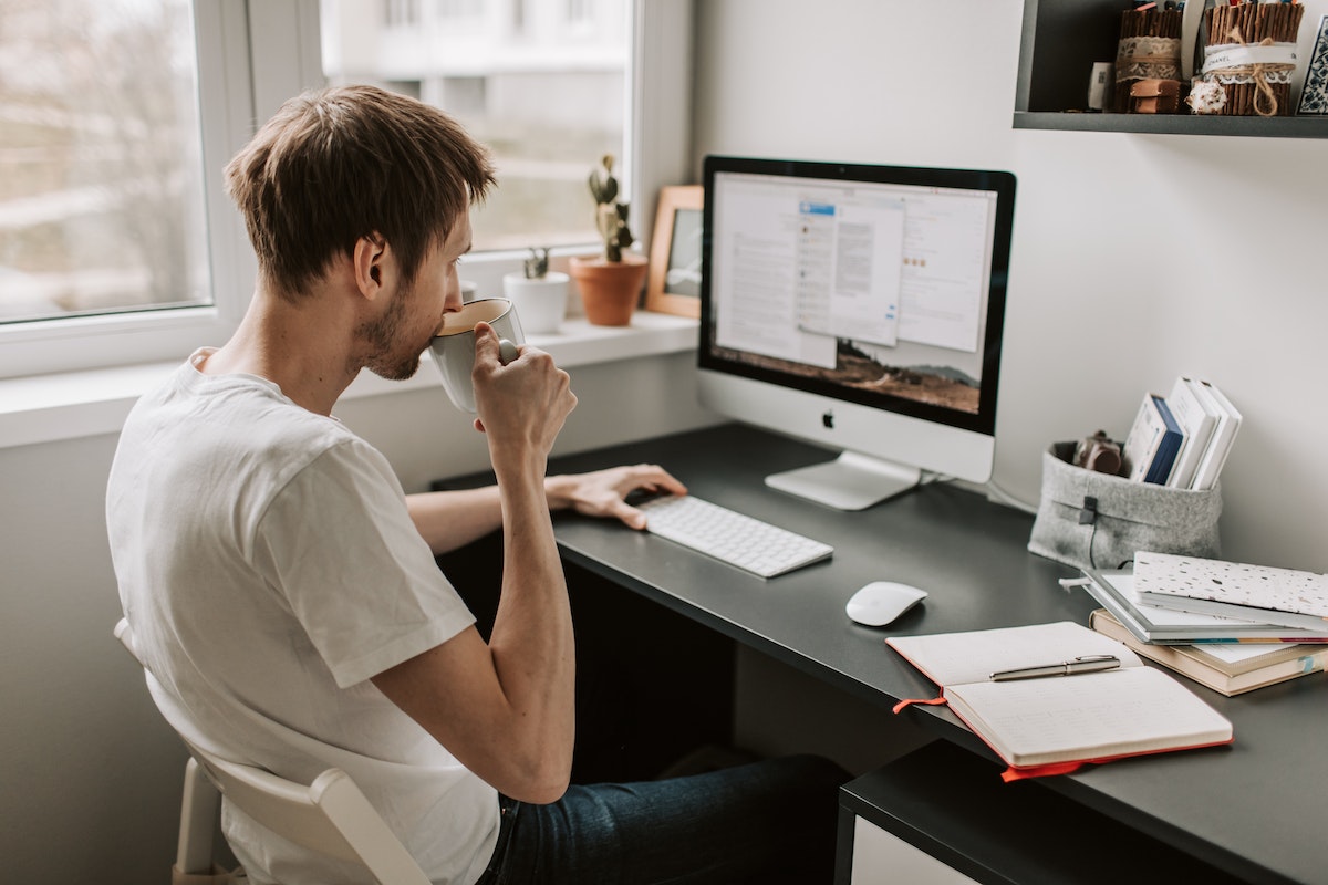 Man in white shirt sitting at a desk drinking coffee in front of a Mac computer