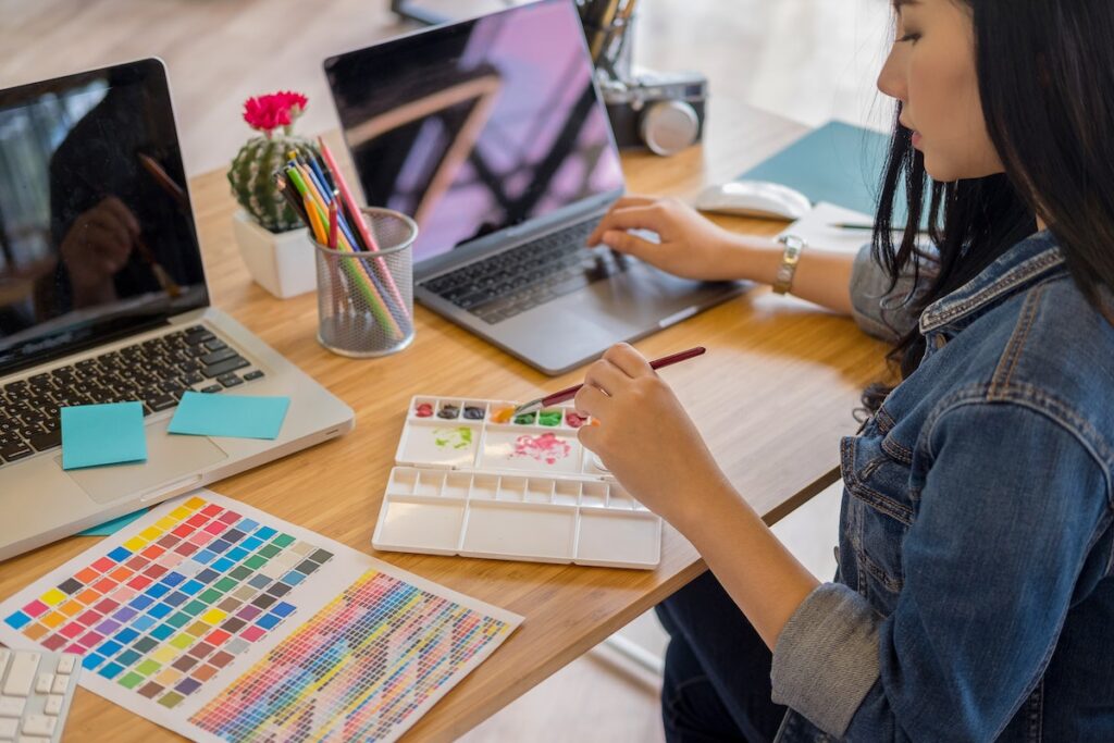 Woman holding paintbrush while working on laptop, creating art, color palette next to her, sittin on a wooden desk, stationary and cactus with red flowers in front of her.