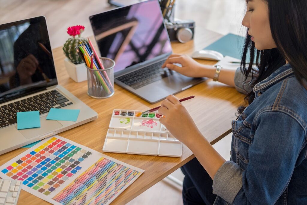 Woman holding paintbrush while working on laptop, creating art, color palette next to her, sittin on a wooden desk, stationary and cactus with red flowers in front of her.
