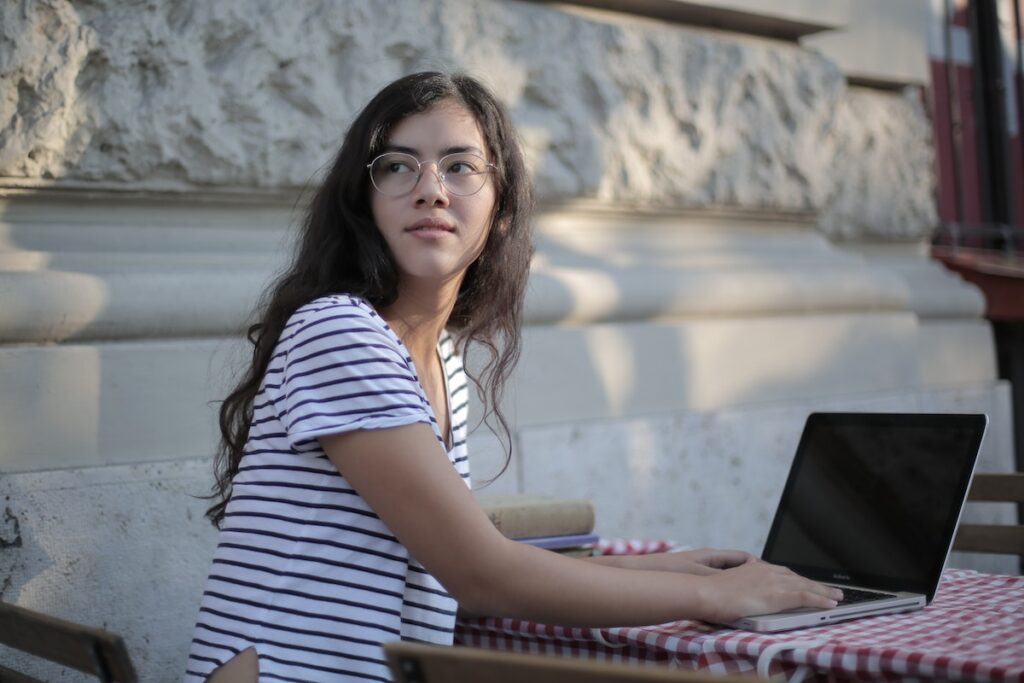 Woman in a striped t-shirt sitting at an outside table with her laptop, looking behind her
